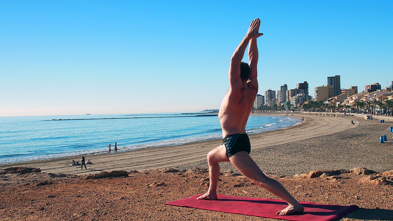A person in Warrior I on a beach, front knee bent and both arms raised overhead