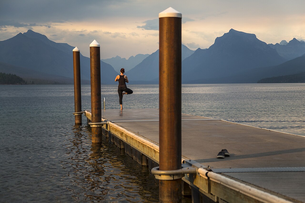 A person balancing on one leg in Tree Pose on a wooden pier beside a mountain lake